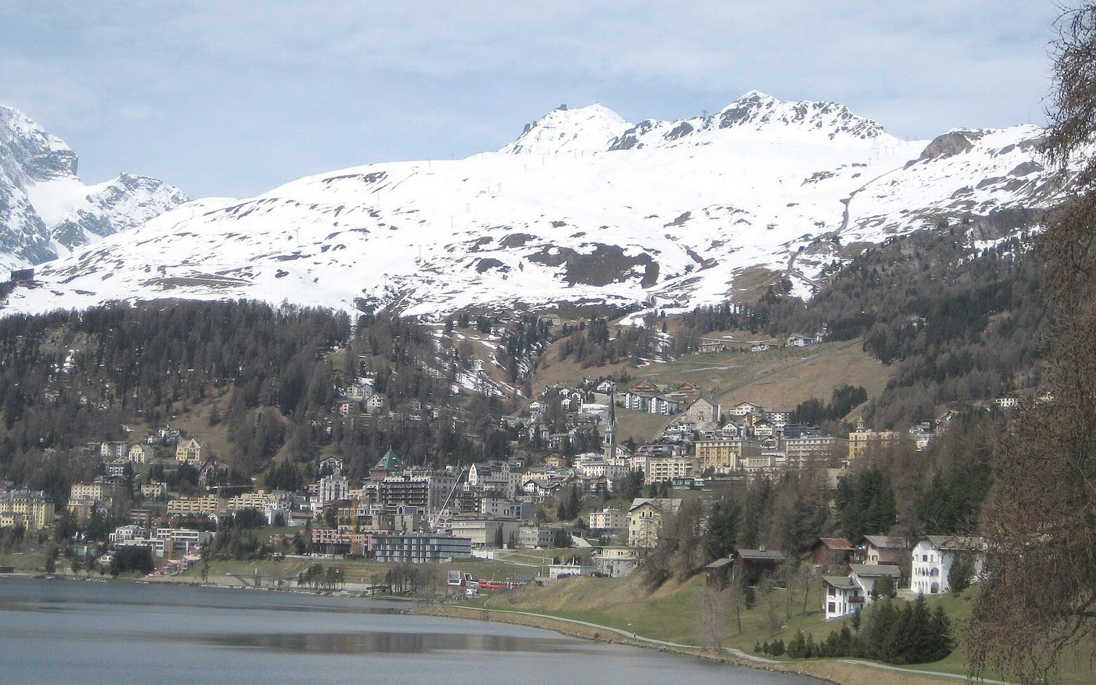 Alpine town of St. Moritz nestled by a lake beneath snow-capped mountains and forested slopes